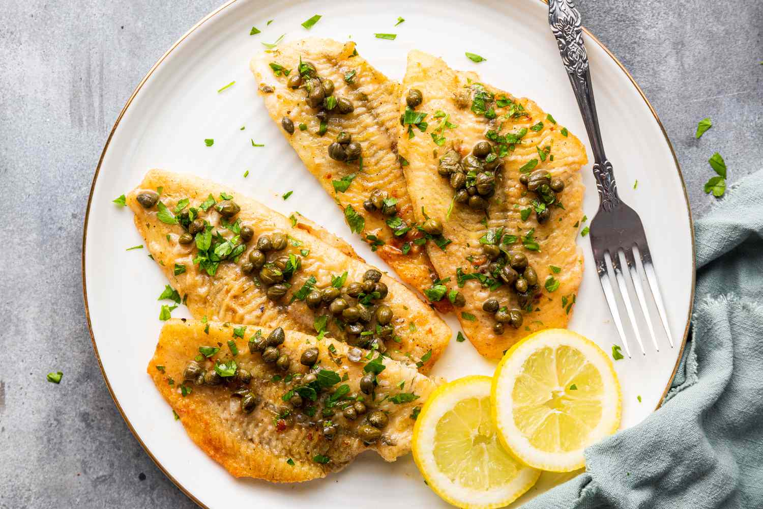 Sole piccata on white plate with a fork and slices of lemon, garnished with chopped parsley
