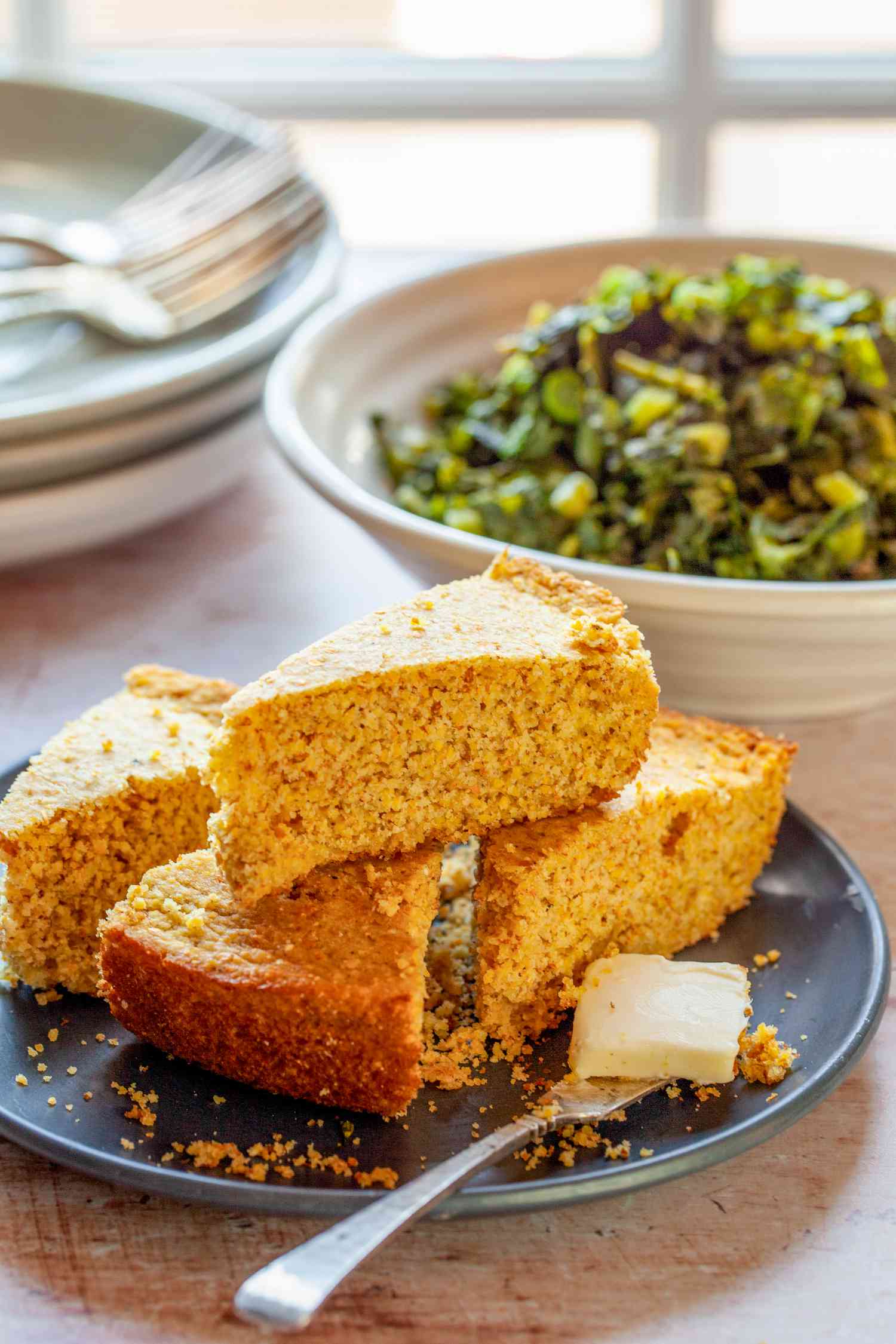 Slices of Southern Cornbread Stacked on a Plate and in the Background, a Bowl of Collard Greens and a Stack of Plates