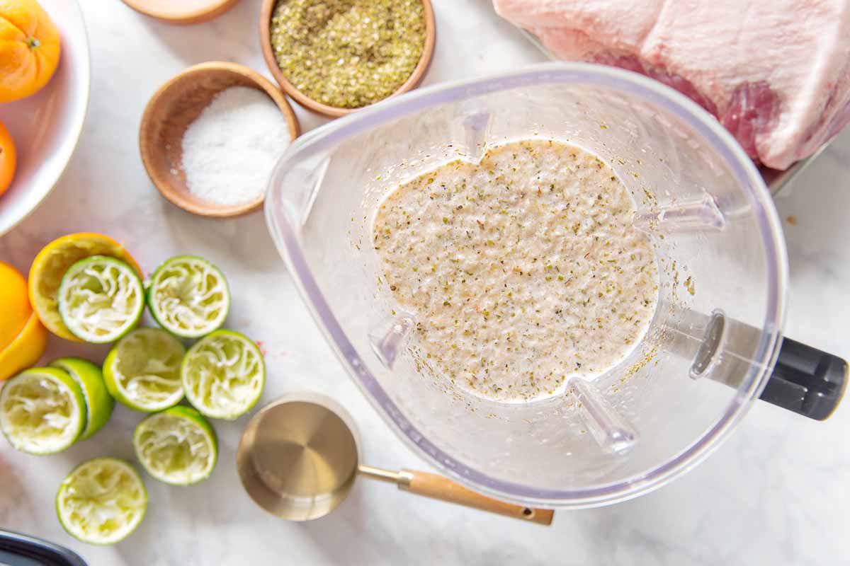 Top view of blender container with pureed marinade inside. Juiced limes, measuring cup, salt, spices and pork roast are around the pitcher and on a marble background.