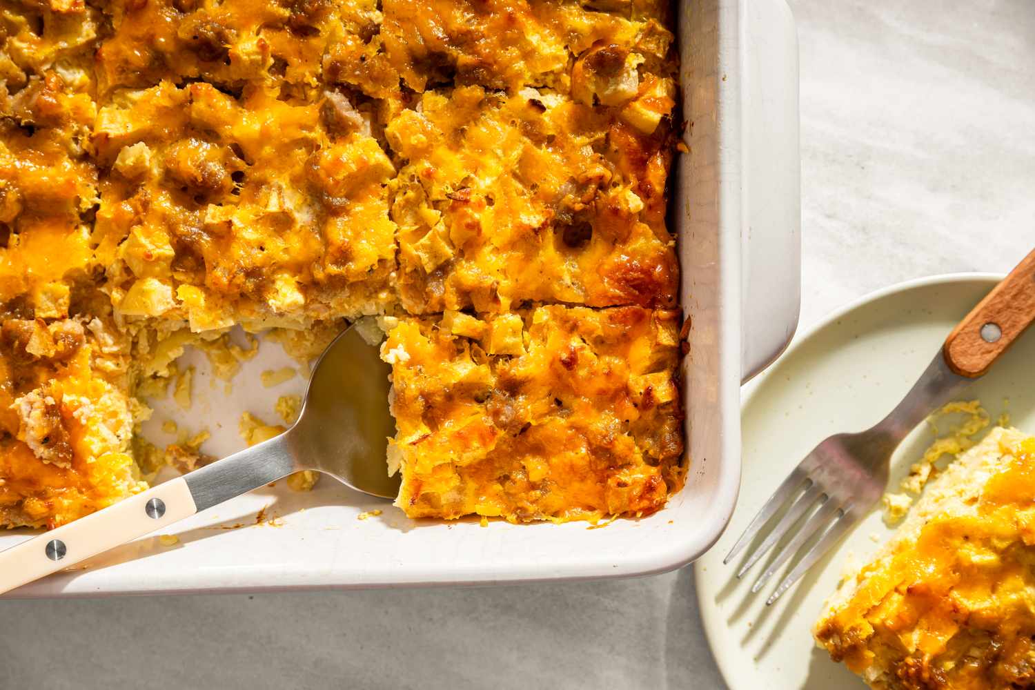Cheesy apple sausage casserole with a serving spatula in a baking dish alongside a plated portion on a small plate with a fork