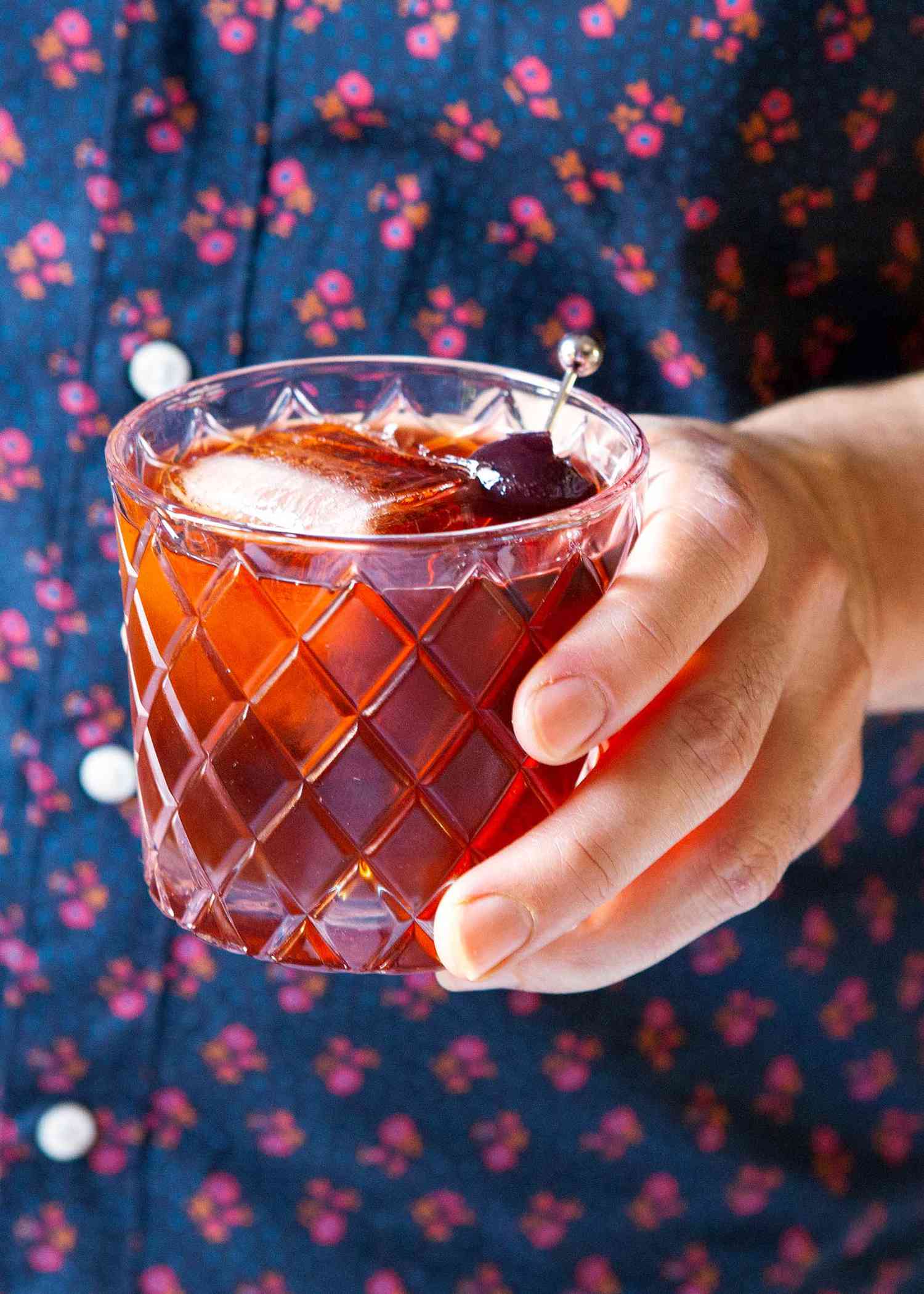Vieux carre with square ice cubes and a cherry garnish are held in the hand of a person.