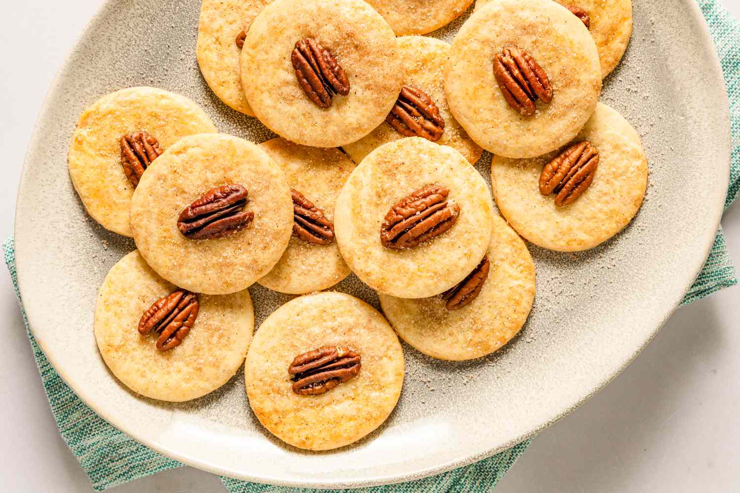 Overhead view of a beige platter of sand tart cookies topped with a pecan halves resting on a green napkin