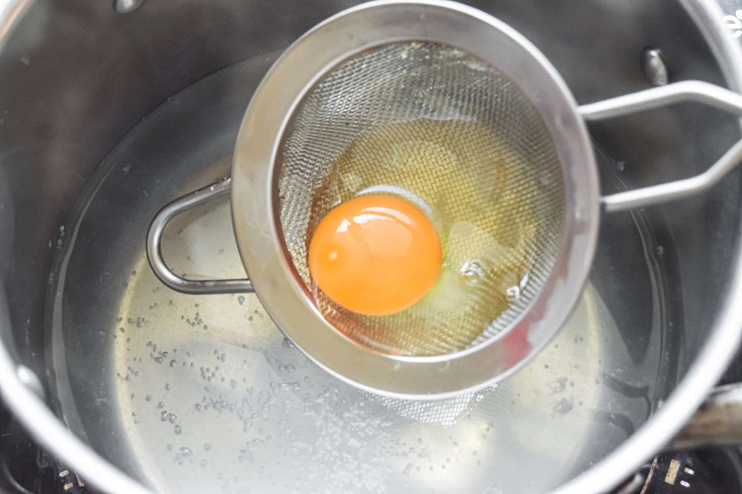 Adding a cracked egg in a sifter to a pot of simmering water 
