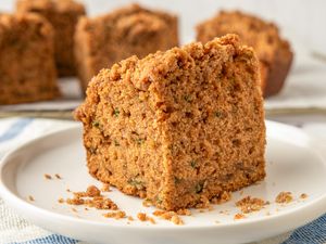 A slice of zucchini coffee cake on a white plate with a crumb topping and background showing additional slices on a cooling rack
