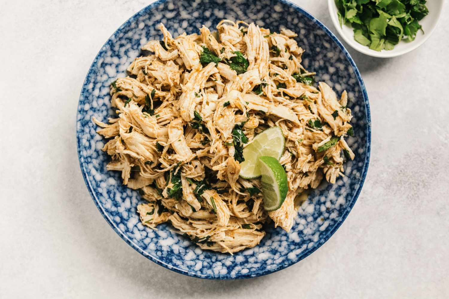 Shredded chicken garnished with herbs and lime served in a blue bowl with a side of chopped parsley in a small dish