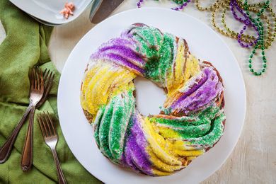 Top view of a homemade king cake on a white plate. Forks and a green linen are to the left. A small plastic baby figurine is above as well as Mardi Gras beads