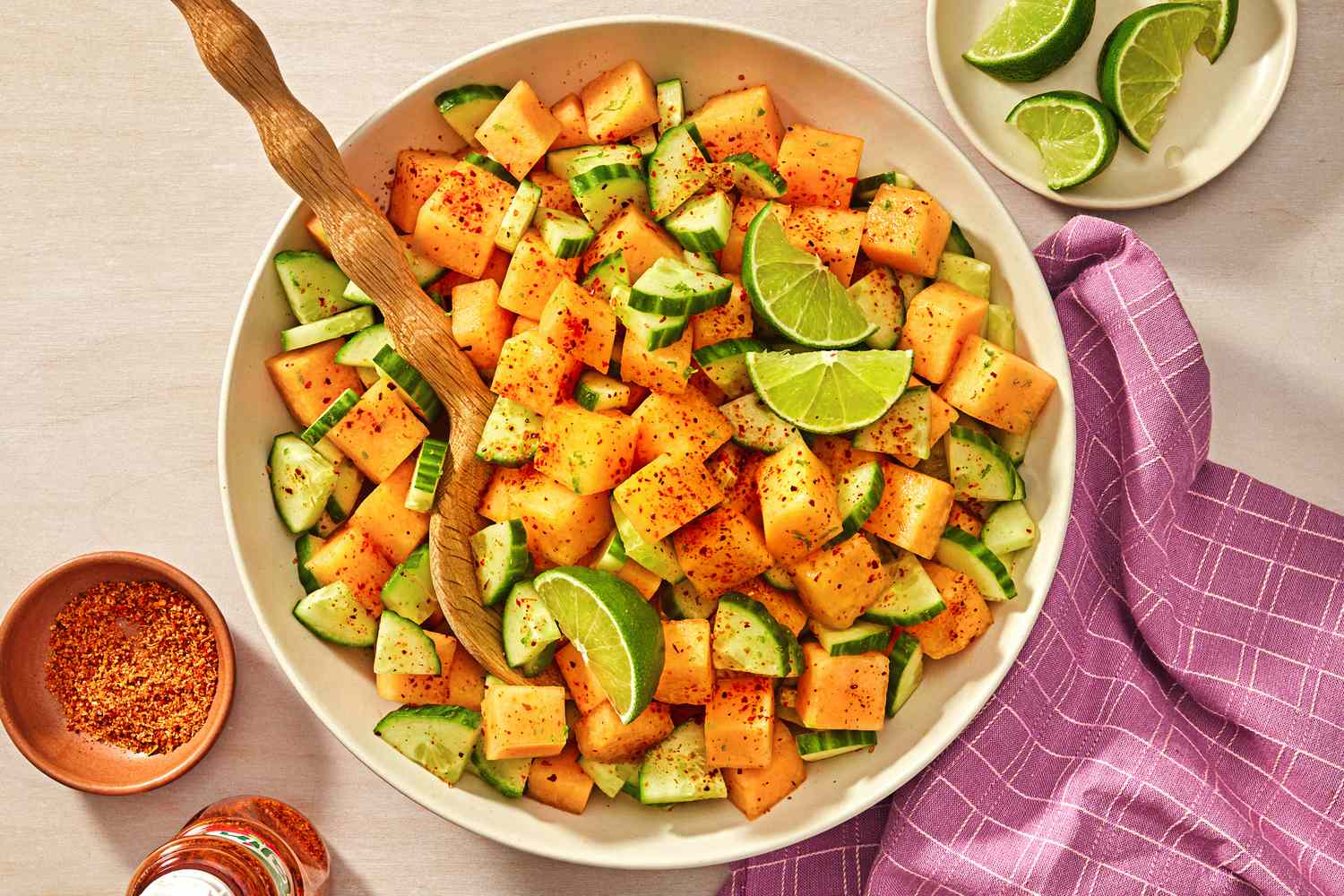 A bowl of cantaloupe and cucumber slices with lime wedges and a sprinkle of Tajn placed on a table with a wooden spoon and a purple cloth