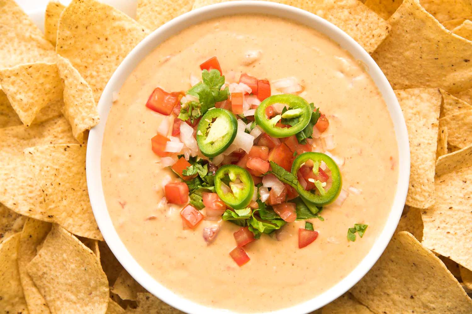 A bowl of queso dip garnished with tomatoes onions cilantro and jalapeo slices surrounded by tortilla chips