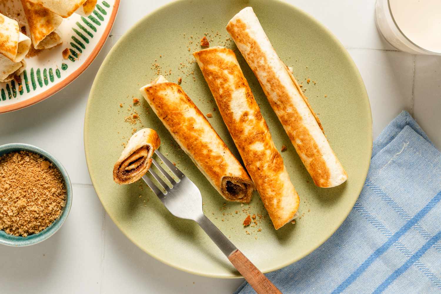 Four cinnamon sugar roll-ups on a plate with a fork, sugar in a bowl nearby, arranged on a fabric surface