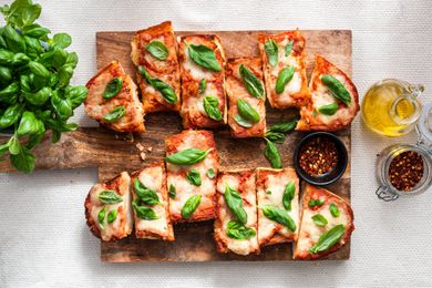 Sliced French Bread Pizza Topped With Fresh Basil (Unsliced) and a Small Bowl of Crushed Peppers on a Wooden Platter and on the Counter Next to It, an Olive Oil Dispenser, a Jar With More Crushed Red Peppers, and a Bushel of Fresh Basil