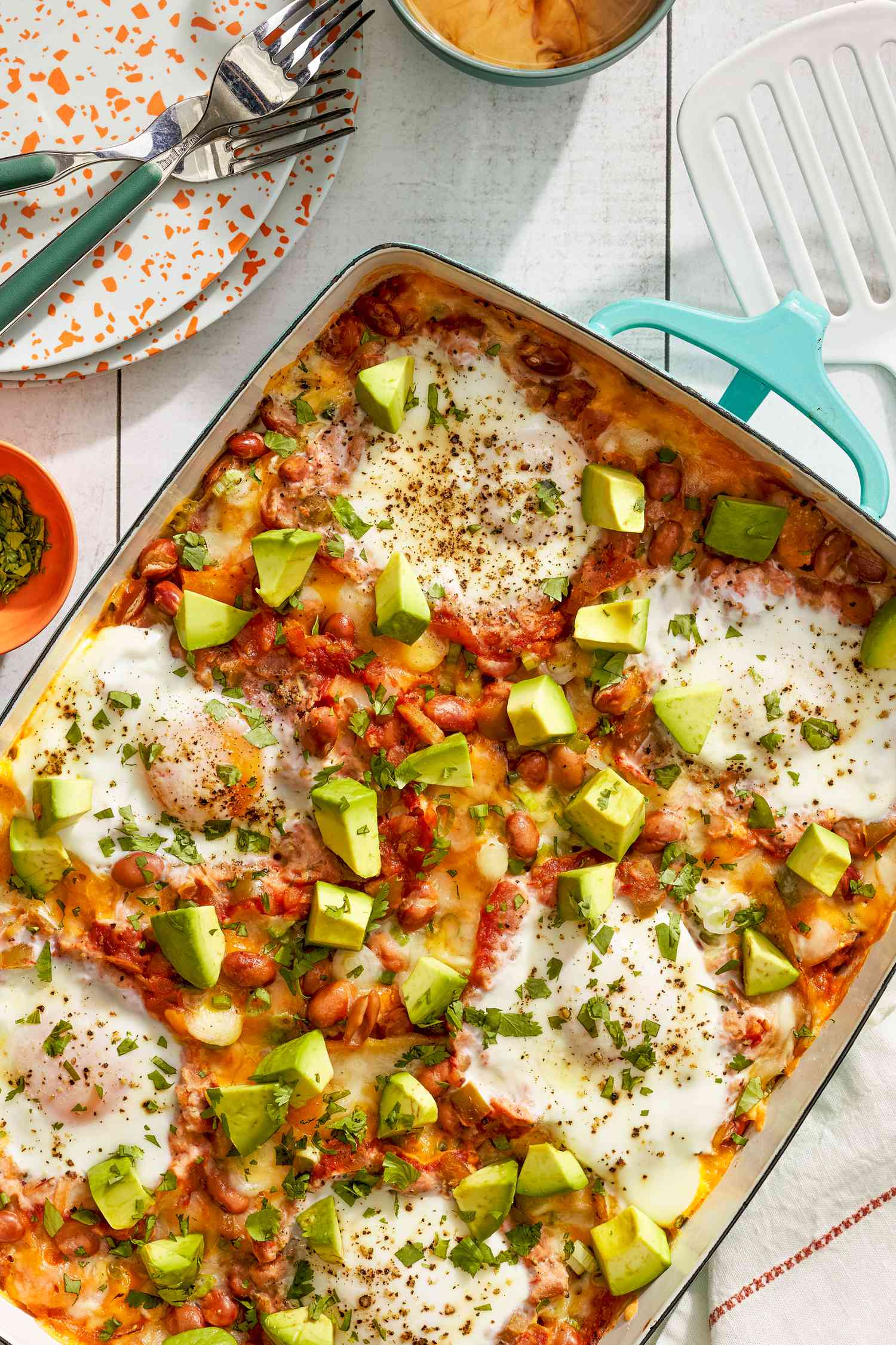 Huevos rancheros casserole at a table setting with a stack of plates and utensils, a cup of coffee, a small bowl of herbs, and a spatula