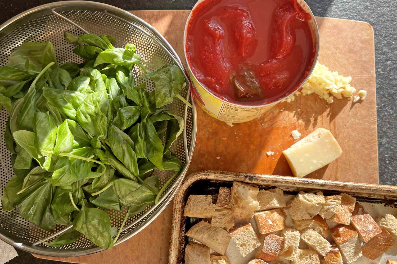 Ingredients including fresh basil canned whole tomatoes parmesan cheese garlic and bread cubes on a cutting board