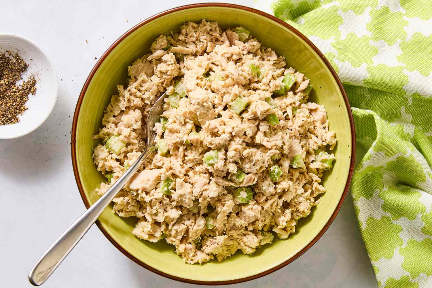 A bowl of tuna salad with diced celery, a silver spoon placed within, next to a napkin and a small dish of spices