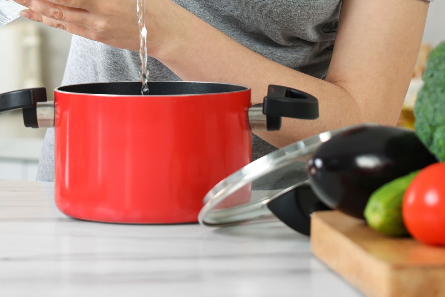 Person pouring water into a red cooking pot with vegetables and a lid nearby