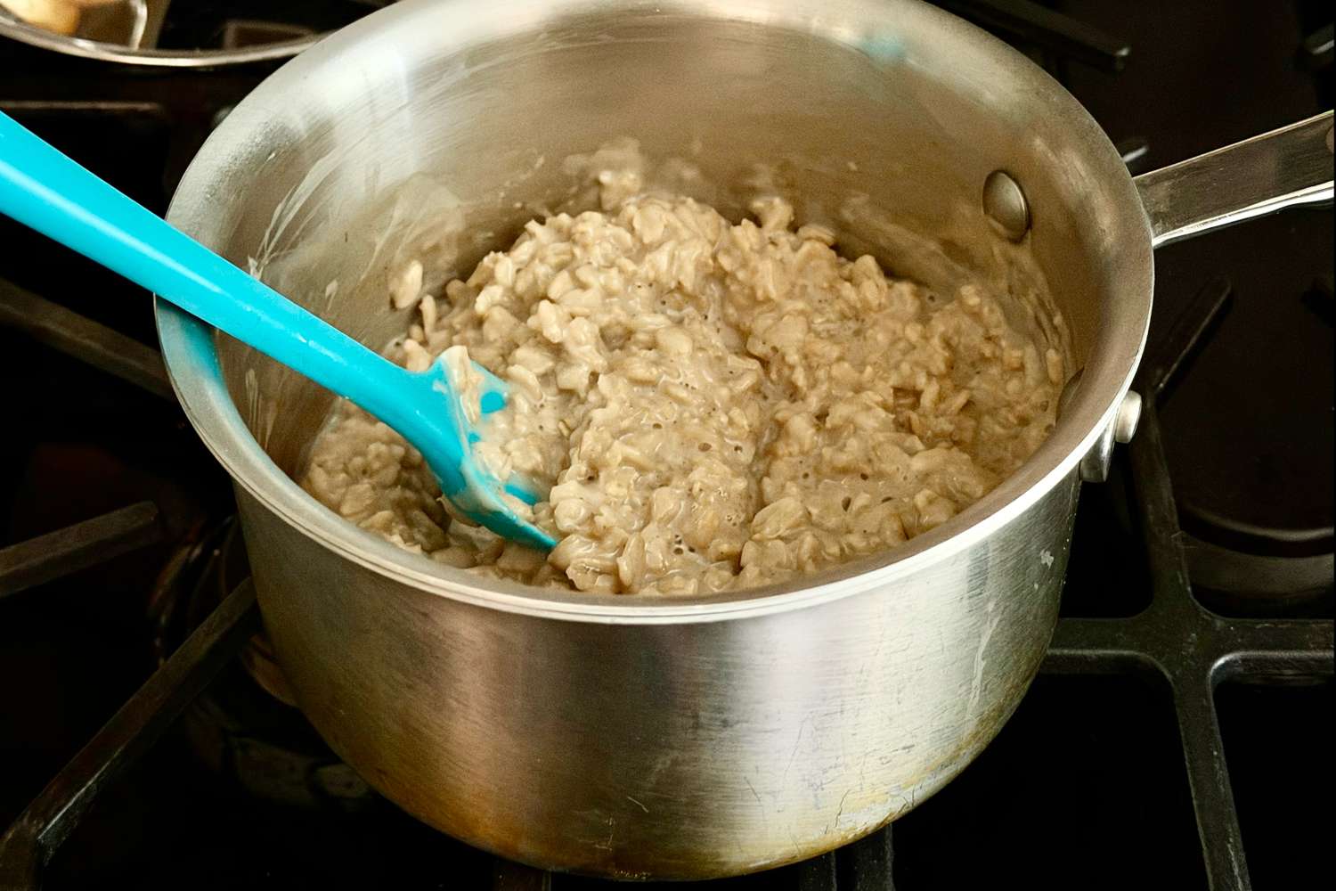 A saucepan filled with cooked oatmeal on a stovetop stirred with a blue spatula