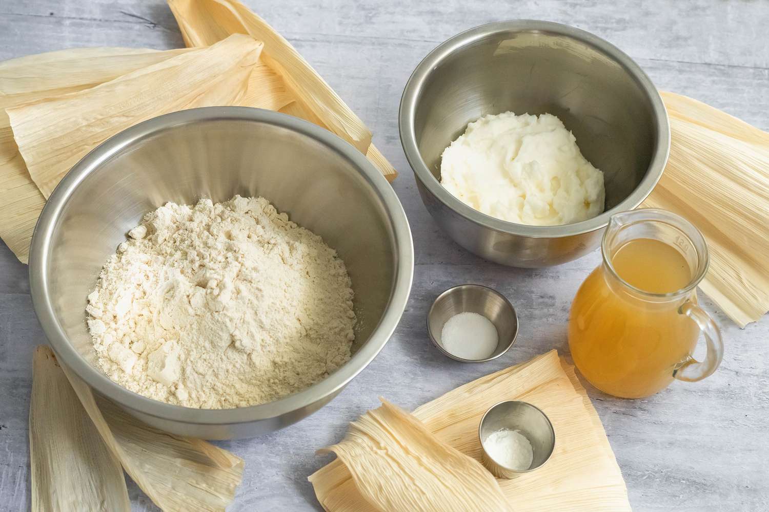 Masa (Tamales Dough) Ingredients (Bowl of Lard, Bowl of Masa Harina, Bowl of Salt, Bowl of Baking Powder, and Pitcher of Chicken Stock All Sitting on Corn Husks)