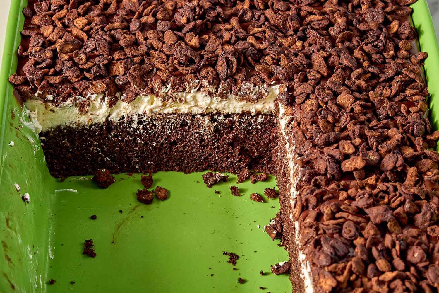 A chocolate and cream layered cake in a green baking dish with a portion removed topped with chocolate chips