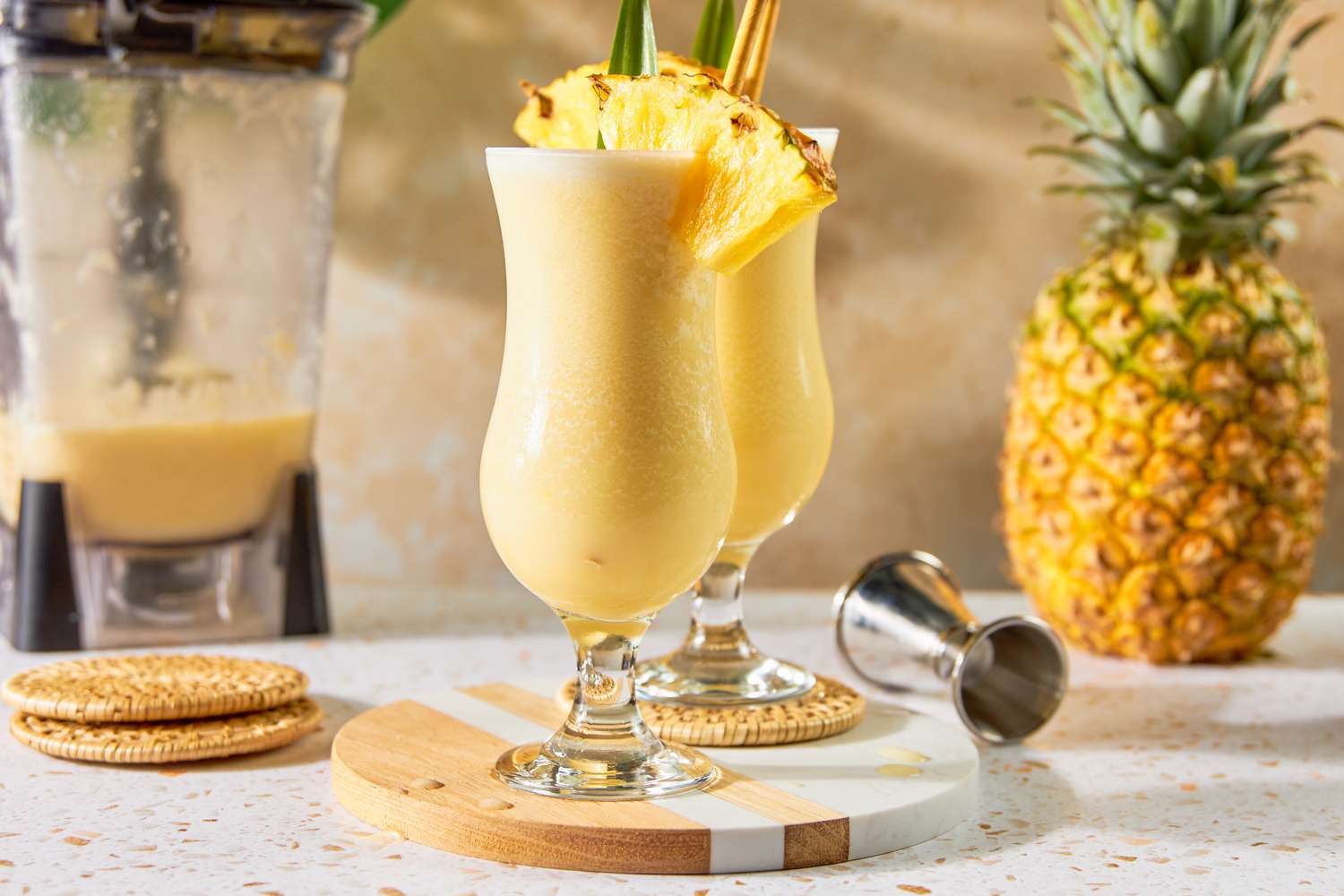Two Glasses of PiÃ±a Colada With a Pineapple Wedge, Pineapple Leaf, and a Straw on a Marble and Wood Circular Serving Tray, and in the Background, an Uncut Pineapple, a Jigger, and a Blender With Some Leftover PiÃ±a Colada