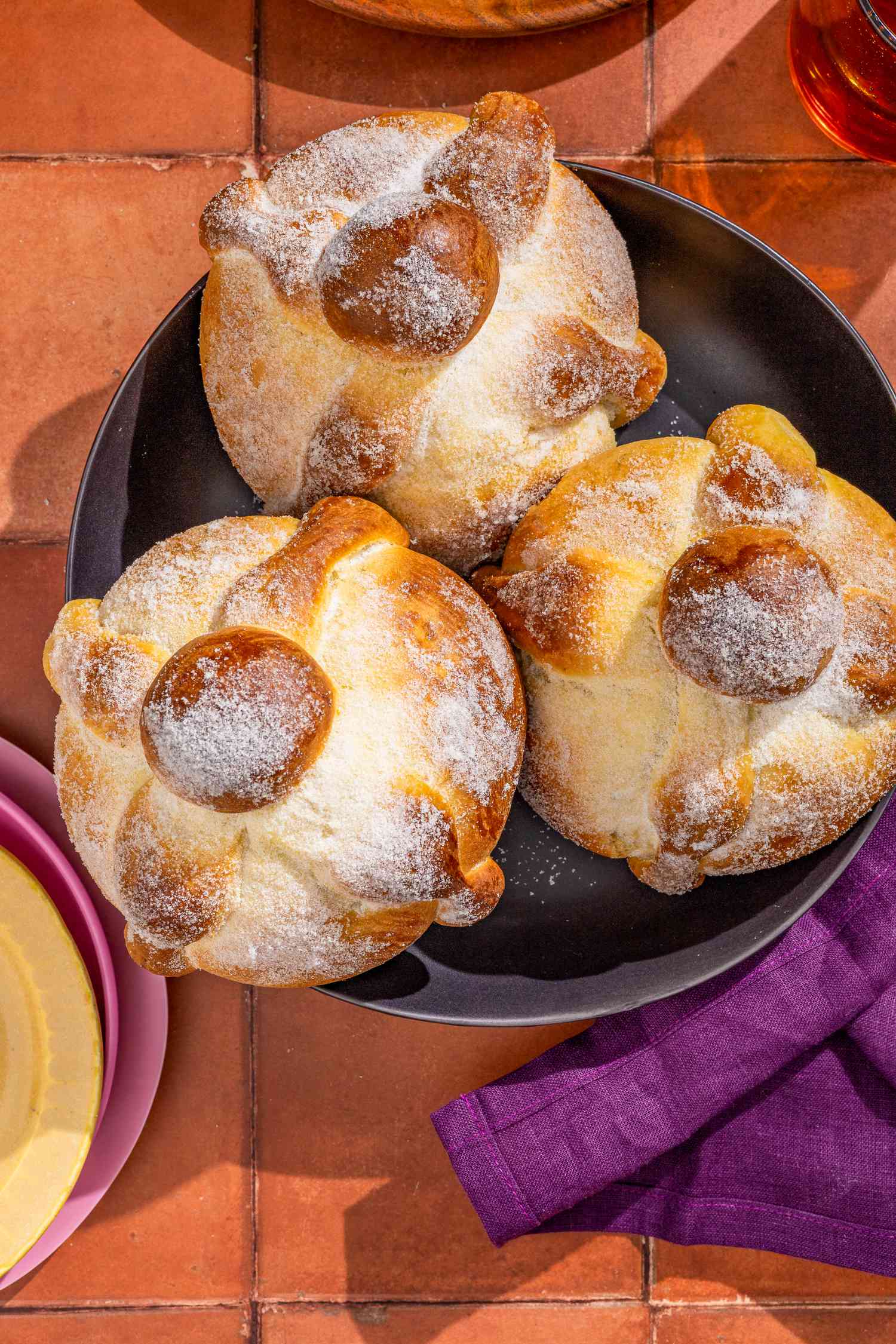 Pan de muerto in a bowl next to a stack of plates, a purple kitchen towel, and glasses of water, all on a brick surface
