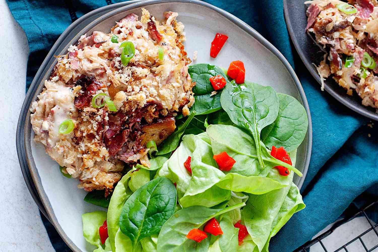 Close up view of a ceramic plate with a scoop of easy reuben casserole and side salad with diced red peppers. A second plate with reuben casserole is partially in view to the right. A dark teal linen is underneath the plates.