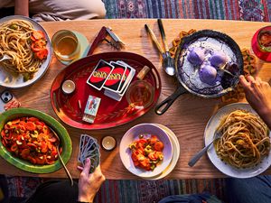 Coffee table filled with food including pasta, carrot salad, and ube skillet cookie, Hands are digging into the food and playing uno