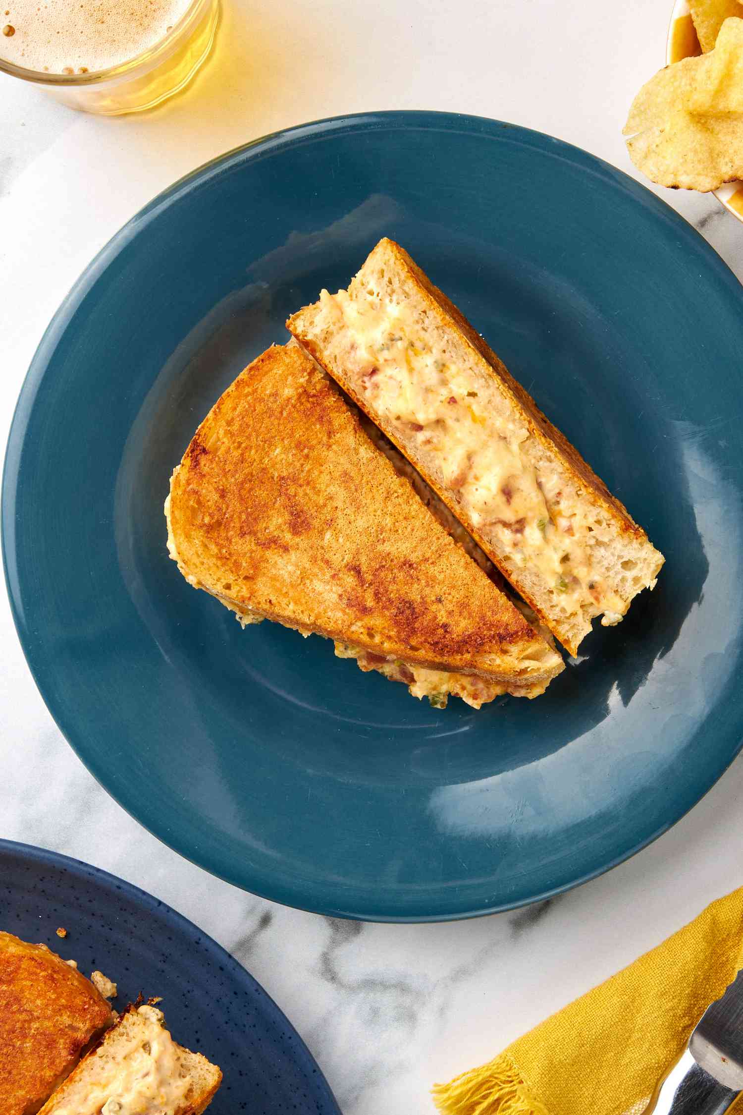 Plate with jalapeno popper grilled cheese (cut in half diagonally and showcasing a cross-section), and in the surroudnings, a bowl of kettle chips, a drink, a serrated knife on a yelllow kitchen towel, and another plate of grilled cheese