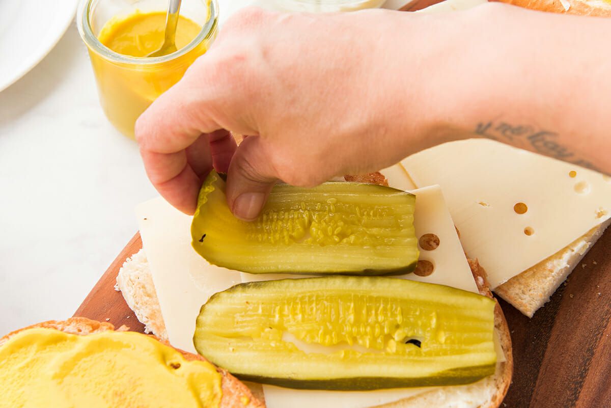 Hand is placing sliced pickles on top of sliced cheese and french bread. One top of the sandwich is covered with mustard and another part of bread has sliced swiss cheese on it. A glass container of yellow mustard is visible in the top left corner.