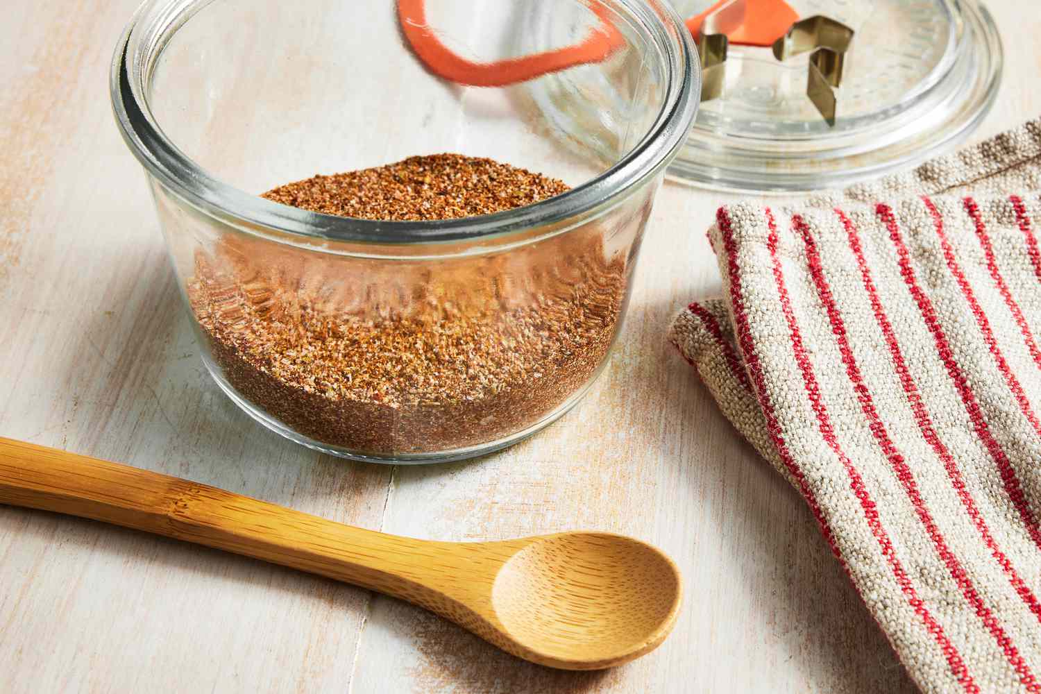Fajita Seasoning in a Glass Jar, and in the Surroundings, a Wooden Spoon, a Red and White Stripped Kitchen Towel, and a Glass Lid for the Jar 