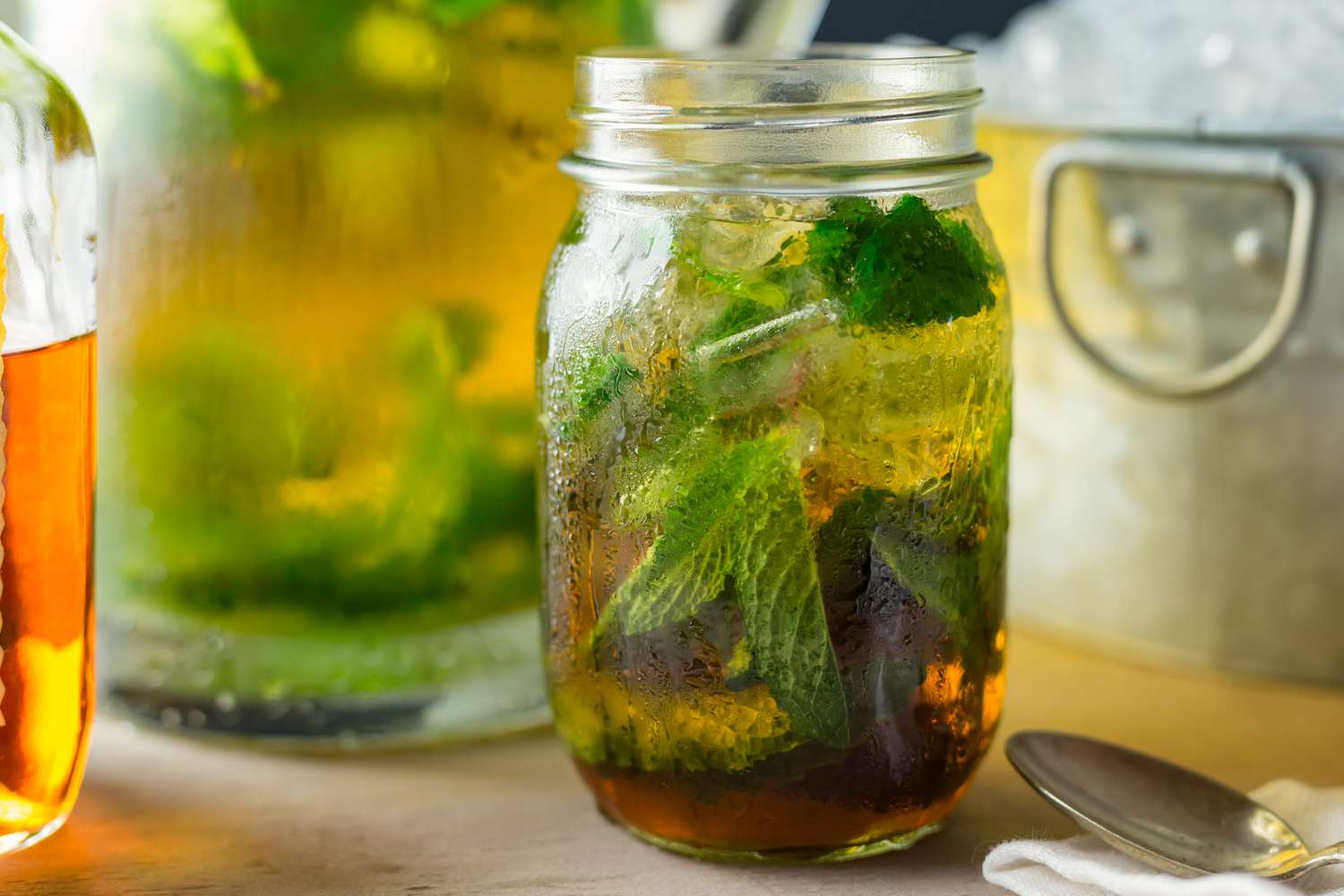 Side view of a mason jar of mint julep, ice and mint leaves with a tub of ice and pitcher in the background