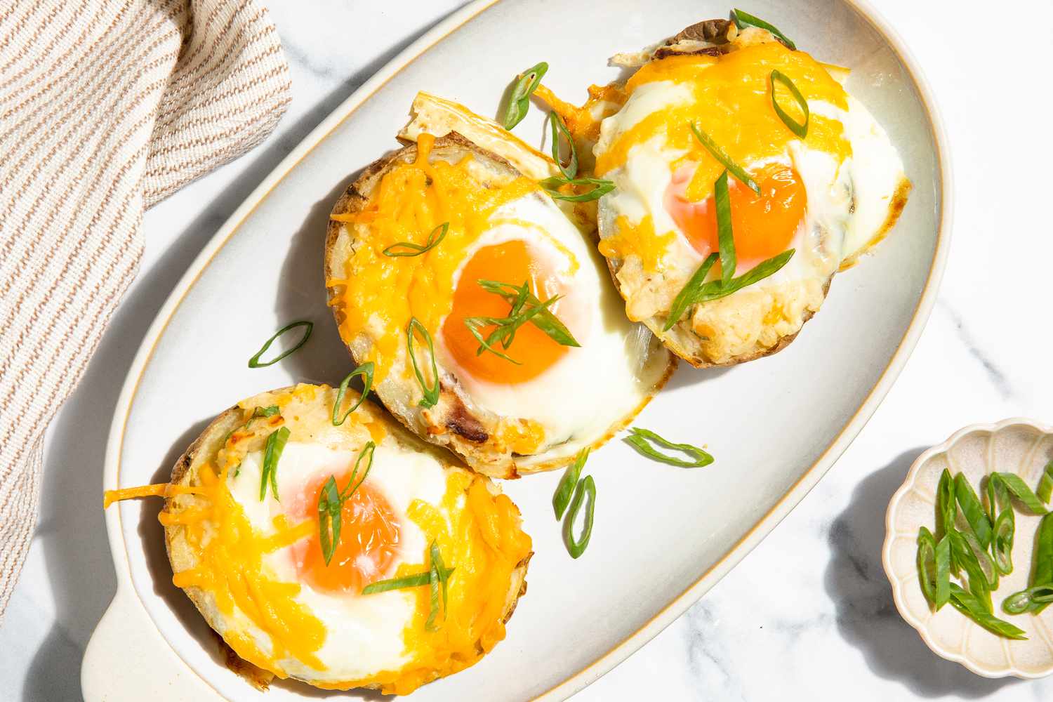 Overhead view of a platter with three loaded potatoes next a small bowl of green onions and a cloth napkin all on a marble countertop