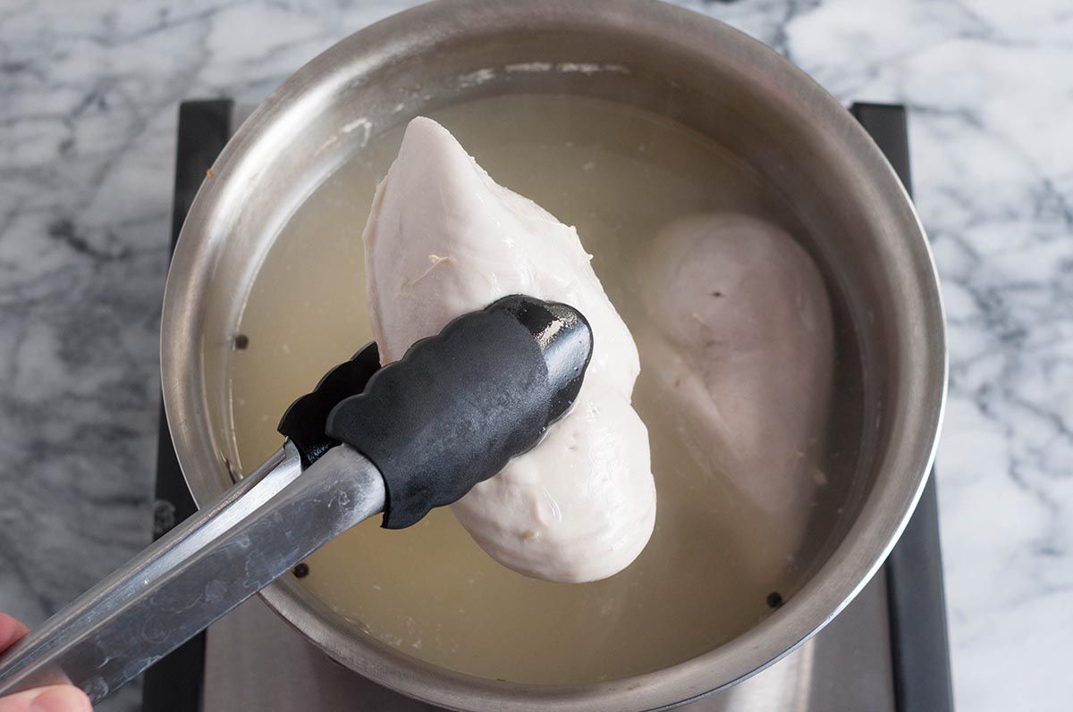 Tongs pulling a cooked chicken breast out of a saucepan of broth