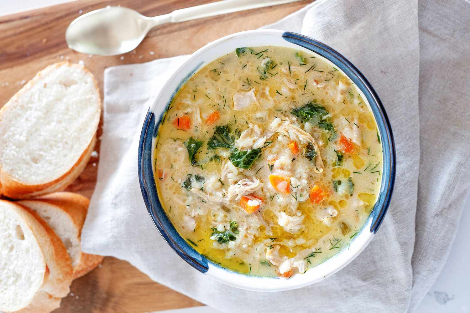 Overhead view of Yasmin Khan’s Lemon Chicken Soup along with sliced bread and a spoon on a cutting board.