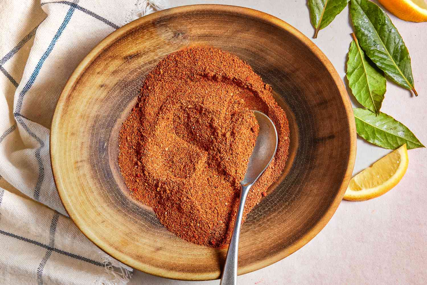Bowl of Old Bay Seasoning With a a Spoon, Sitting on a White Kitchen Towel Next to Lemon Leaves and Lemon Wedges