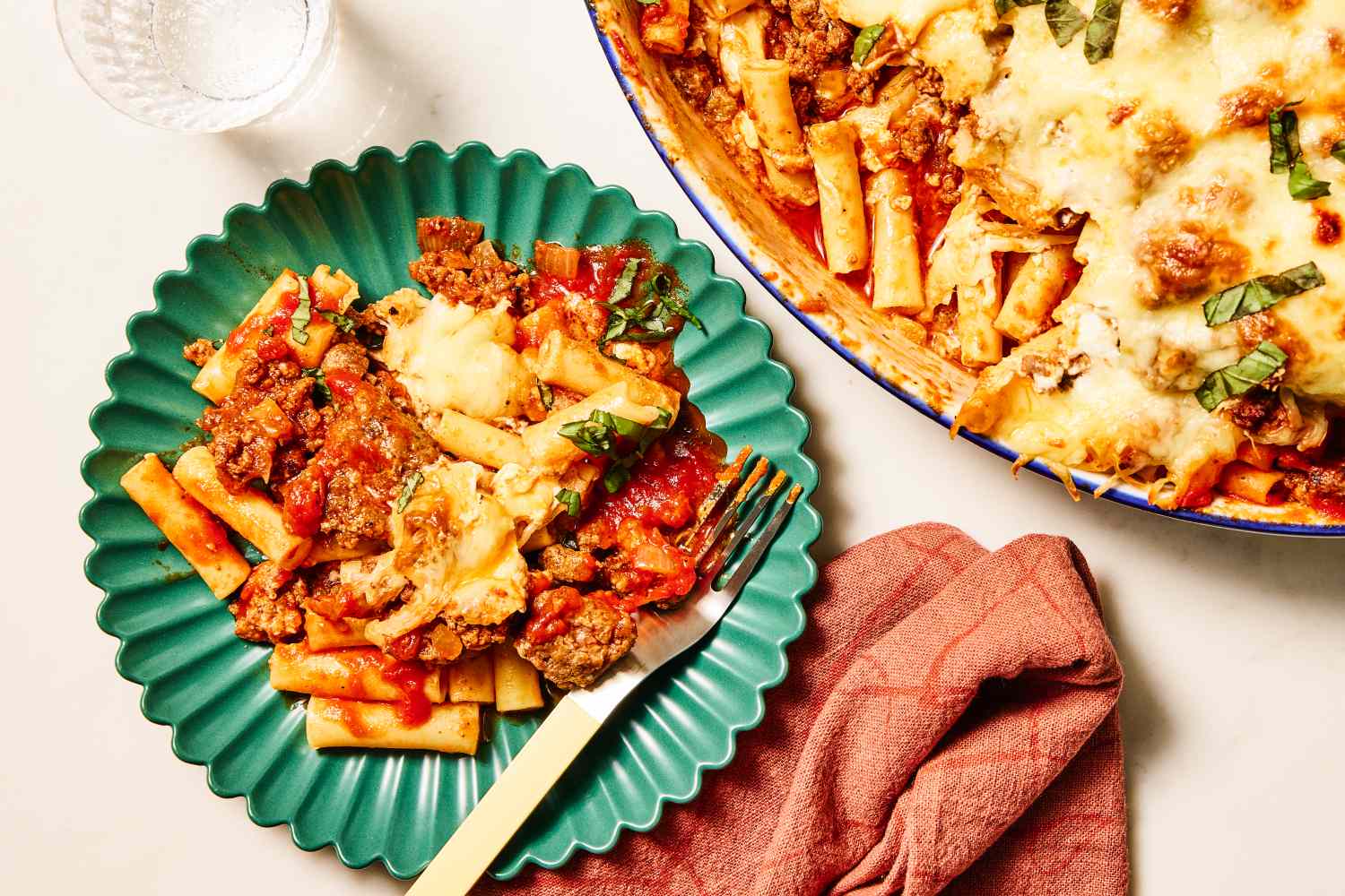 A serving of baked ziti with cheese and herbs on a plate fork on the plate next to a dish with the remaining pasta
