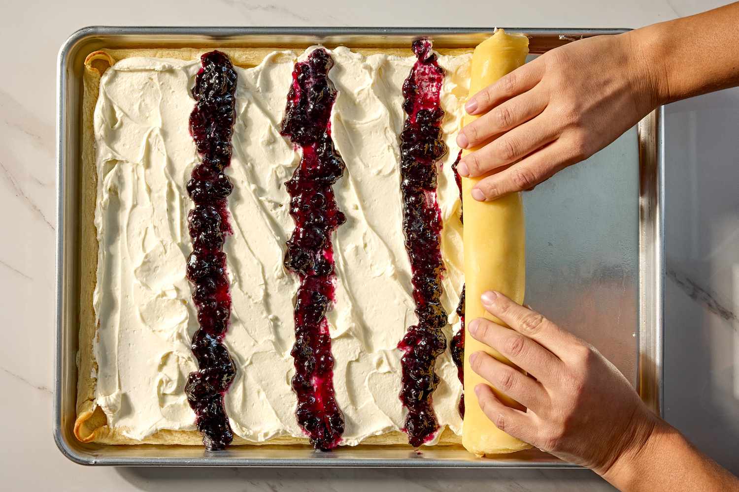 Hands rolling up a crepe layer with cream and fruit filling on a baking tray