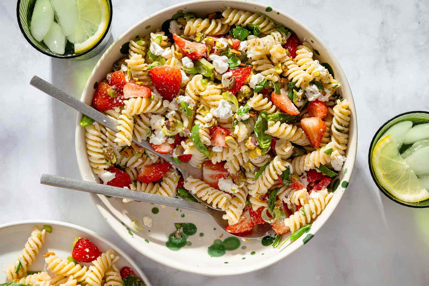A bowl of rotini pasta salad with strawberries and greens on a table with drinks in glasses