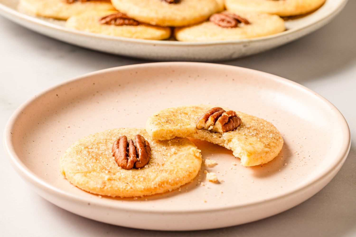 Closeup view of a pink plate with two sand tart cookies topped with a pecan halves and one cookie has a bite taken out of it