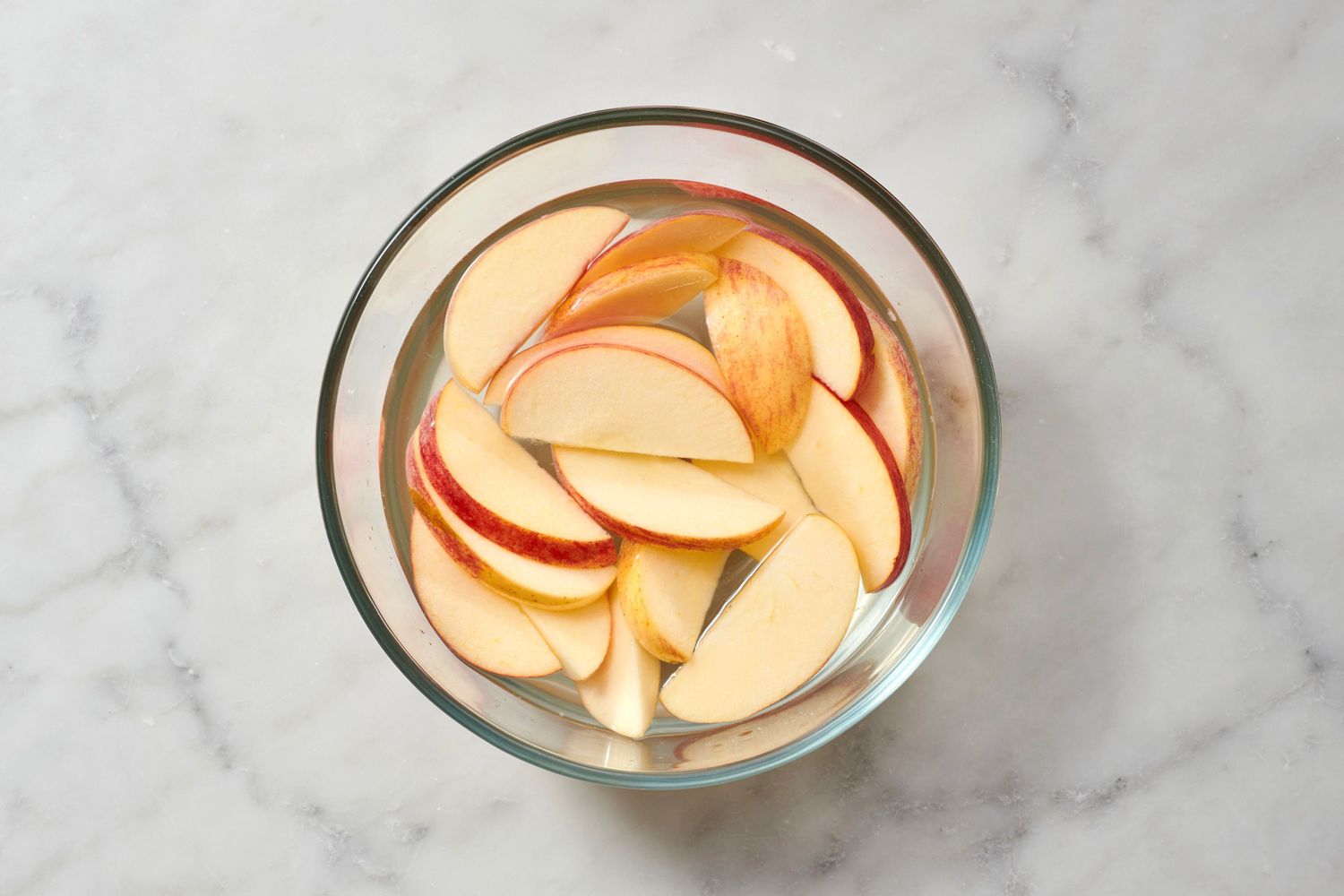 Sliced apples submerged in a clear bowl of liquid on a marble surface