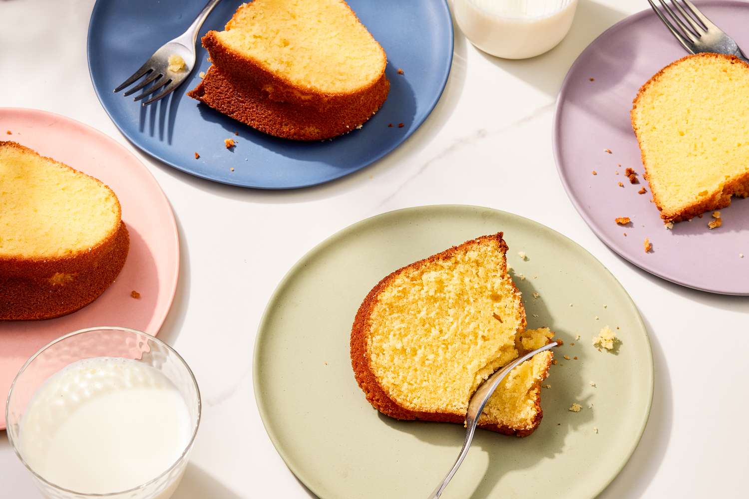Overhead view of 4 plates with slices of pound cake and forks next to glasses of milk on a marble countertop
