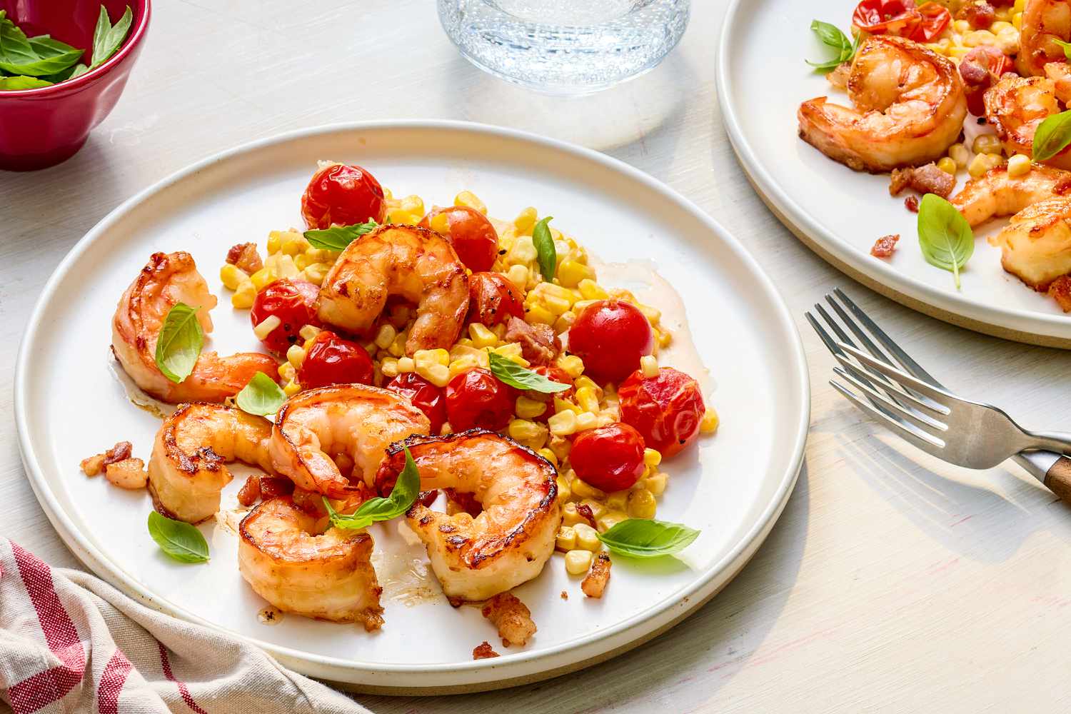 Plate of creamy skillet shrimp with corn, pancetta, and tomatoes at a table setting with another serving on a plate, a glass of water, utensils and a white and red table napkin on the counter, and a bowl of basil