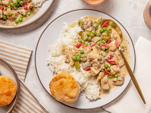 Serving of chicken a la king on a plate with rice and a biscuit at a table setting with another serving on a plate, a bowl of biscuits, glasses of wine, and table napkins