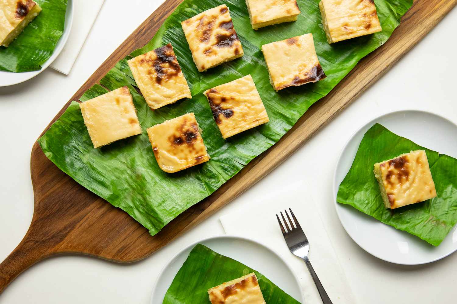 Aerial Shot: Cassava Cake Slices on a Banana Leaf Covered Wooden Tray, and Individual Slices of Cake on Banana Leaf Covered Plates
