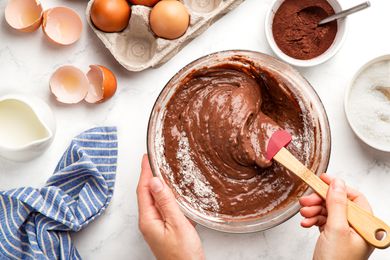 someone mixing chocolate cake batter in a bowl at a table with a carton of eggs, a glass of milk, a bowl of cocoa powder, and a bowl of flour
