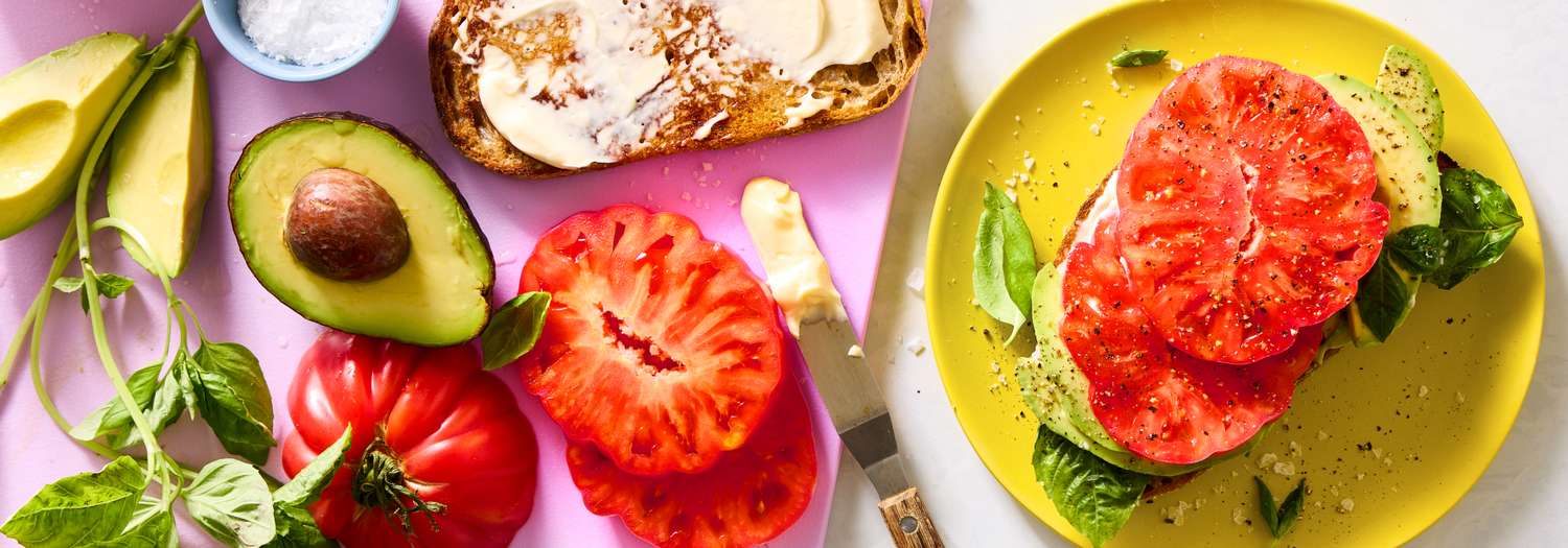 Ingredients including sliced tomatoes, avocado halves, toasted bread with spread, and basil leaves arranged for a meal
