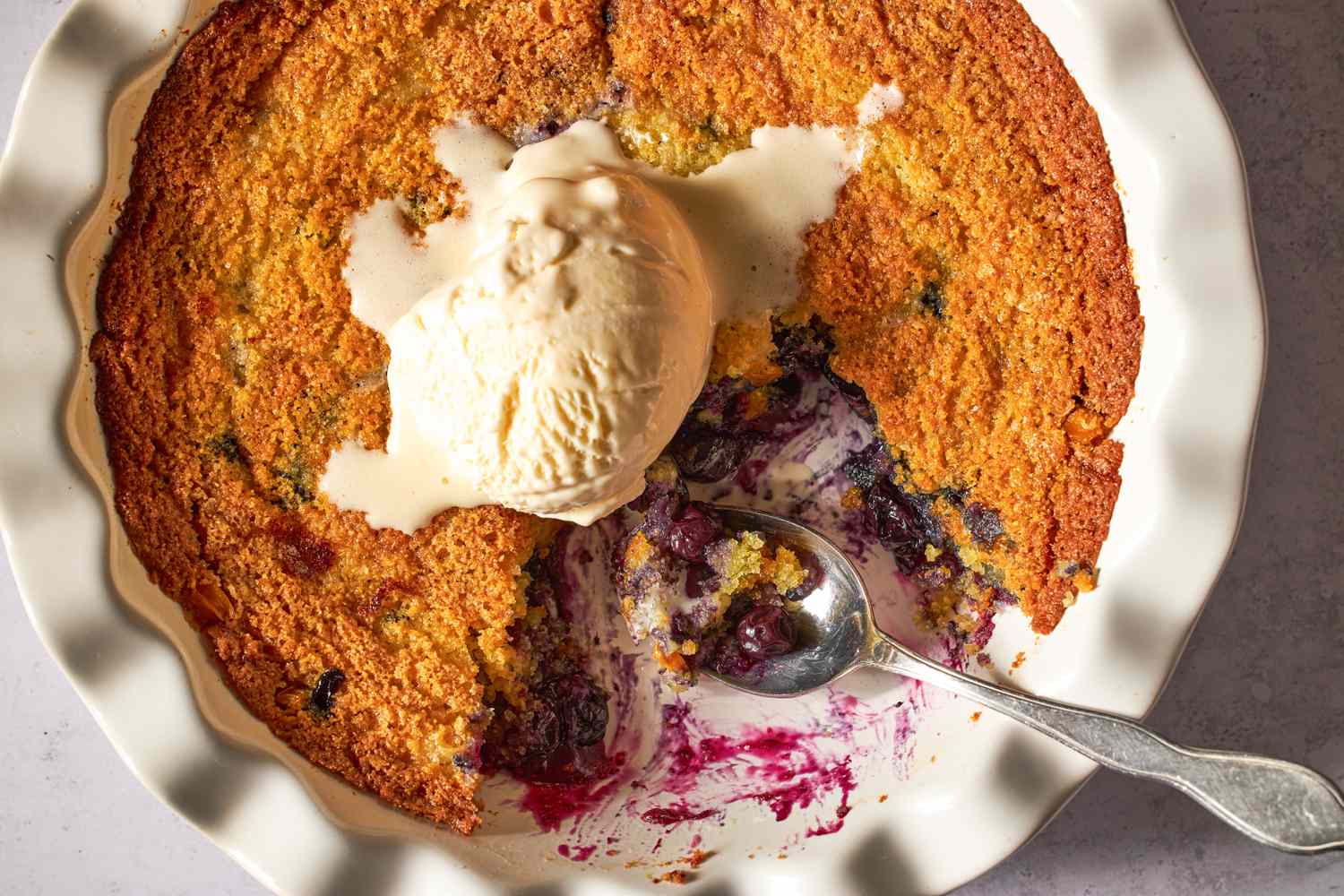 Blueberry corn cake with a scoop of vanilla ice cream on top in a fluted baking dish, served with a spoon