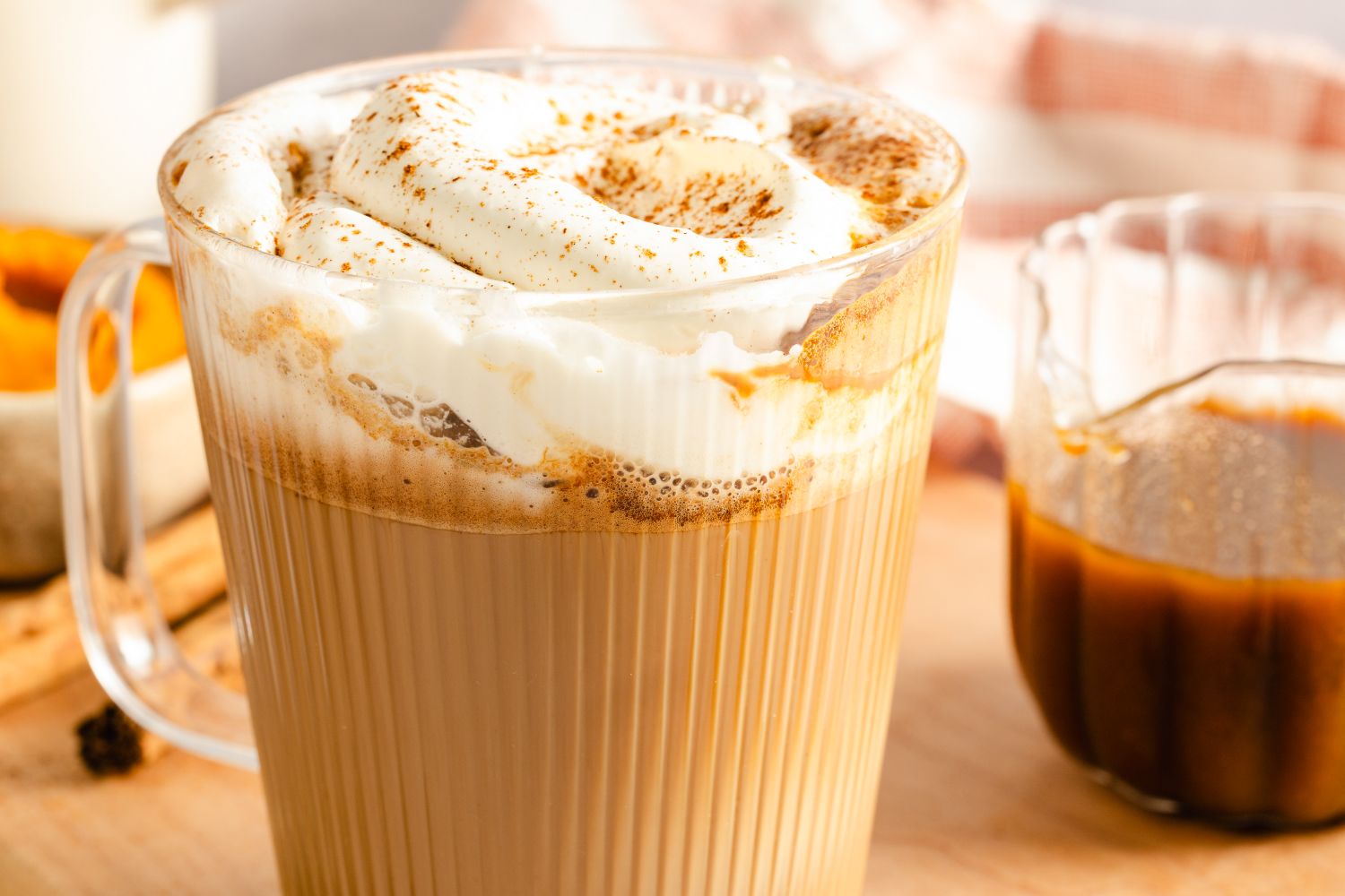 Glass mug of pumpkin spice latte topped with whipped cream and a jar of spiced syrup nearby