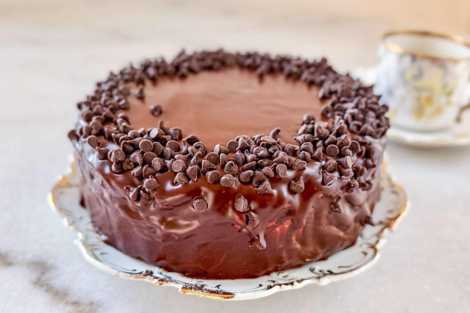 A chocolate cake topped with chocolate chips on a decorative plate with a teacup in the background