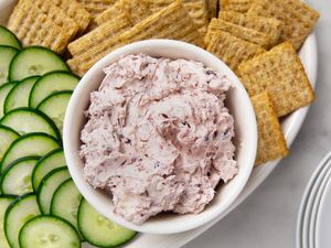 A bowl of cream cheese spread surrounded by crackers and sliced cucumbers on a platter