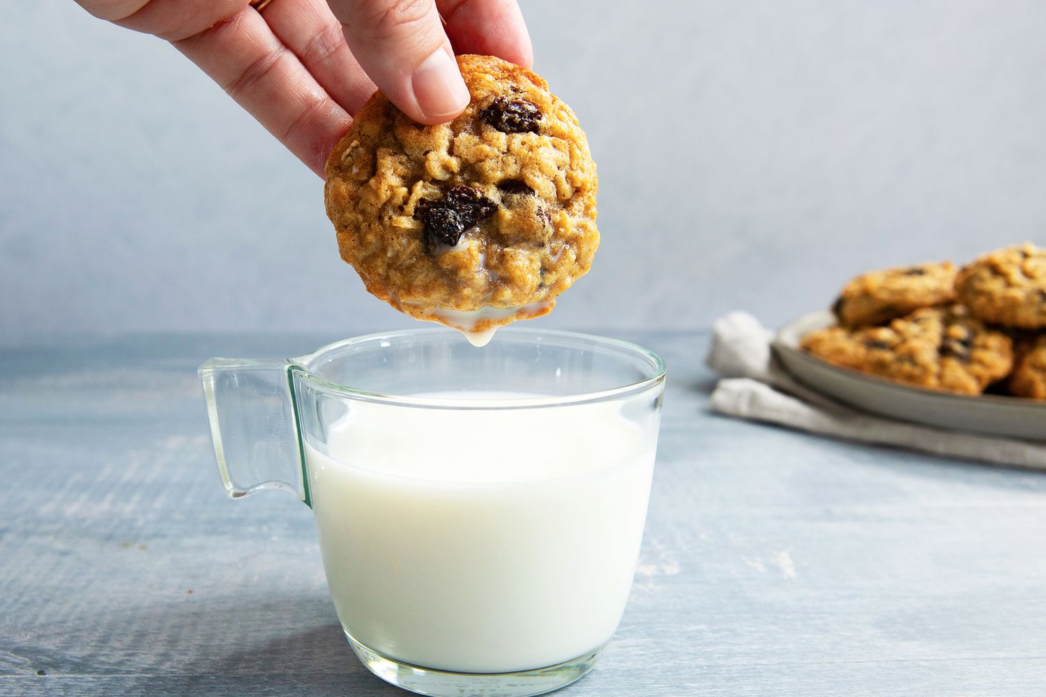 A person dipping an oatmeal raisin cookie into a glass of milk additional cookies on a plate in the background