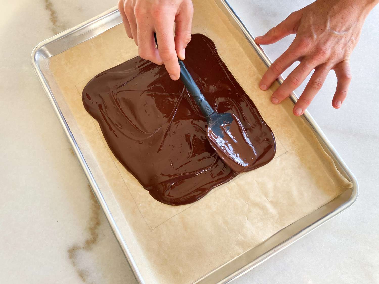 A hand holding a silicone spatula spreading melted chocolate on a parchment paper-lined sheet pan